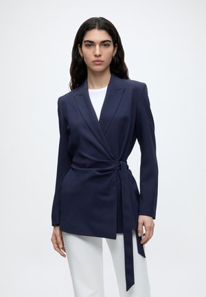 Vrouw met lang donker haar die een marineblauwe overslagblazer draagt, gestrikt in de taille, een wit overhemd erboven en een witte broek tegen een effen achtergrond.