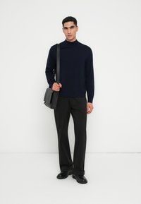 Marineblauer Pullover mit hohem Kragen, kombiniert mit schwarzen Hosen und einer schwarzen Umhängetasche. Das Modell steht vor einem neutralen Hintergrund.