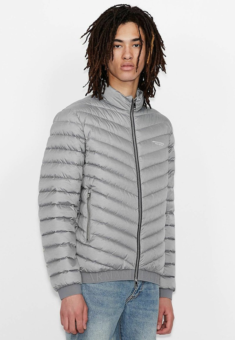 Grå pufferjakke med chevron-quiltet design, høj krave, frontlynlås og sidelommer. Funktioner ribbede manchetter og mærket bæltedel.