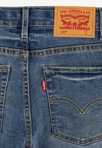 Pantalones vaqueros de mezclilla azul con costuras naranjas, que presentan un parche de marca de cuero, una etiqueta con un tab rojo y un diseño clásico de bolsillo trasero. Modelo 510.