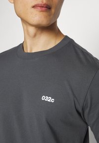 T-shirt gris à col rond en coton doux, arborant un logo blanc 032c sur la poitrine. Design simple avec des manches courtes et une coupe décontractée.