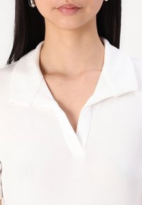 Bílý polo tričko s krátkým rukávem a sklopeným límcem, který má hladkou, měkkou strukturu. Čisté linie a minimalistický design.