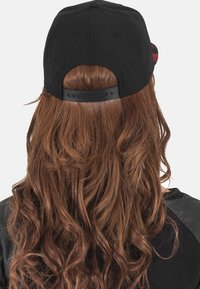 Schwarze Baseballmütze mit verstellbarem Riemen hinten, von hinten betrachtet, vor einem schlichten weißen Hintergrund. Lange lockige braune Haare sind sichtbar.