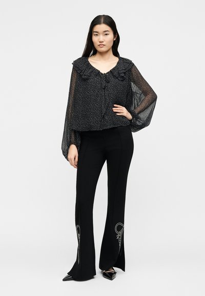 Γυναίκα που φορά διαφανές μαύρο πουκάμισο με λευκές βούλες και βολάν, σε συνδυασμό με μαύρο καμπάνες παντελόνι με λευκή κεντητική κοντά στο τελείωμα.