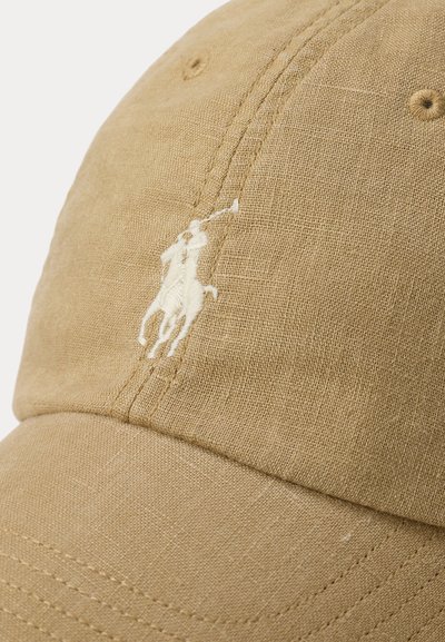 Μπεζ υφασμάτινο καπέλο μπέιζμπολ με κεντημένο λευκό λογότυπο ενός παίκτη πόλο πάνω σε άλογο που κρατάει υψωμένο το ρόπαλο.