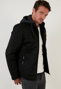 Svart jacka med huva, med texturerade axelpartier och dragkedjefickor. Slät tyg med en snygg design och figursydd passform.