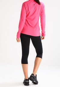 Frau steht mit dem Rücken zur Kamera, trägt ein langärmliges pinkes Oberteil, schwarze knöchellange Leggings und schwarze Sportschuhe.