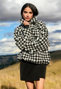 Jachet�ă din polar alb-negru cu model houndstooth, cu guler înalt, manșete elastice și tiv cu șnur, purtată cu o fustă neagră. Textură moale.