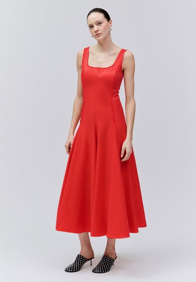 Femme portant une robe midi rouge sans manches à encolure carrée et des mules à talons noires cloutées, debout devant un fond gris uni.