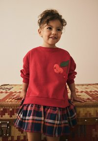 Roter Sweatshirt mit Kirsch-Applikation und einem Rock aus marineblauem Karostoff. Das Outfit zeichnet sich durch weiche Materialien und ein lässiges Design aus.