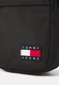Borsa in tessuto nero con una patch rettangolare riportante il testo "TOMMY JEANS" e blocchi colorati in rosso, bianco e blu. Superficie testurizzata.