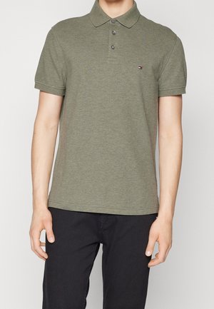 Homme portant un polo vert olive à trois boutons avec un petit logo brodé sur le côté gauche de la poitrine, associé à un pantalon noir.