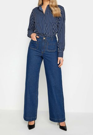 Kobieta ubrana w granatowo-białą koszulę w paski, wpuszczoną do wysokich, szerokich niebieskich dżinsów z kieszeniami na przodzie oraz czarne szpilki.