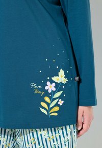 Teal langærmet bluse med blomstermønster og teksten "Flower Breeze", parret med stribede bukser med blomsterdesign, hånden hvilende ved siden af.