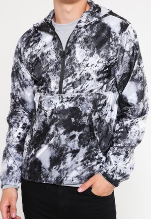 Personne portant une veste à capuche noire et blanche à motif abstrait, avec une fermeture éclair sur le devant et une poche kangourou, main partiellement dans la poche.