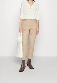 Beżowe spodnie w kant, kremowa koszulka polo i brązowe botki za kostkę. Trzyma dużą białą torbę. Prosty fason z dopasowaną sylwetką i kołnierzykiem.