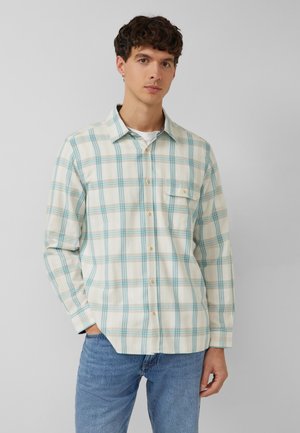 Junger Mann trägt ein cremefarbenes und blaues karierter Hemd mit Brusttasche und hellblauen Jeans, steht vor einem hellgrauen Hintergrund.