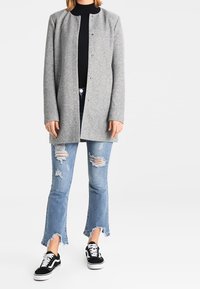 Abrigo gris de mezcla de lana con un diseño recto, frente abierto y cierres de presión. Pantalones vaqueros azules desgastados con dobladillos sin rematar y zapatillas negras.