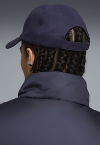 Granatowa czapka z zakrzywionym daszkiem i otwartym tyłem. Noszona z ciemną, ocieplaną kurtką. Charakterystyczne plecione włosy i wyraźna ozdoba na uchu.