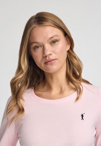 Långärmad rosa tröja med rund halsringning och en liten mörk logotyp på vänster bröst. Mjukt tyg och slät textur.