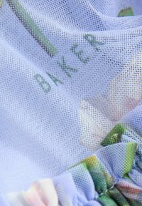 Hellblaues Mesh-Gewebe mit floralem Muster und grünen Rüschenakzenten. Enthält den Text "BAKER" in grüner Stickerei, was ein strukturiertes Erscheinungsbild verleiht.