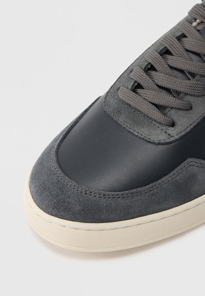 Γκρι αθλητικό παπούτσι με λείο δέρμα και σουέντ υφές, επίπεδα γκρι κορδόνια και μπεζ λαστιχένια σόλα. Καμπυλωτή μύτη και λεπτομέρειες με ραφές.