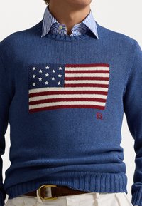 Modrý pletený svetr s výrazným pleteným designem americké vlajky, který obsahuje červené a bílé pruhy a bílé hvězdy. Hladký límec a manžety.