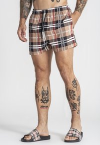 Mann trägt beige, schwarz-weiß-rote karierte Shorts und passende Sandalen, zeigt detaillierte Tattoos an beiden Beinen und Armen.
