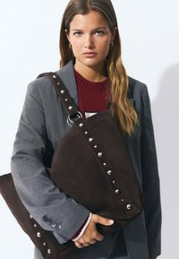 Mujer con cabello largo, vistiendo un blazer gris y un suéter rojo, sostiene un gran bolso marrón oscuro con tachuelas contra un fondo blanco.