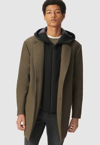 Joven con un abrigo verde oliva sobre una sudadera negra con cremallera y jeans oscuros, de pie contra un fondo liso.