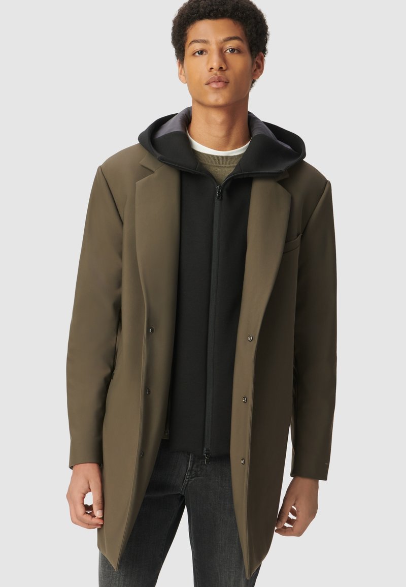 Joven con un abrigo verde oliva sobre una sudadera negra con cremallera y jeans oscuros, de pie contra un fondo liso.