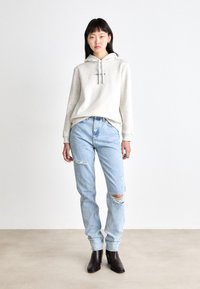 Hoodie cinza claro com um bolso frontal e cordão, combinado com jeans desbotados azuis claros. O modelo usa botas de salto pretas até o tornozelo.