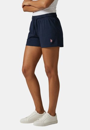 Frau trägt marineblaue Shorts mit Kordelzug und Logo am linken Bein sowie weiße Sneakers, steht mit verschränkten Armen.