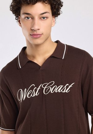Jonge man met krullend haar, die een bruin kort mouw gebreid poloshirt draagt met witte "West Coast" tekst op de voorkant.