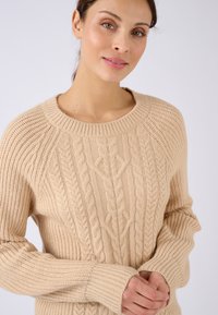 Beige gebreide trui met kabelbreipatronen, een ronde halslijn, lange mouwen en ribgebreide manchetten. Zachte textuur met een relaxte pasvorm.