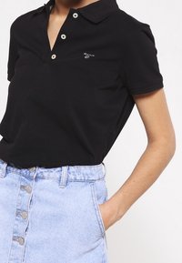 Polo noir avec un col, trois boutons, et un petit logo sur la poitrine, associé à une jupe en denim bleu clair à boutons.