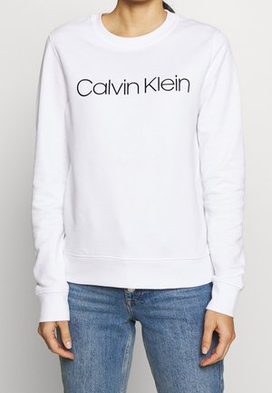 Mujer vestida con un sudadera de manga larga blanca de Calvin Klein y jeans azules, de pie con las manos relajadas a los lados frente a un fondo liso.