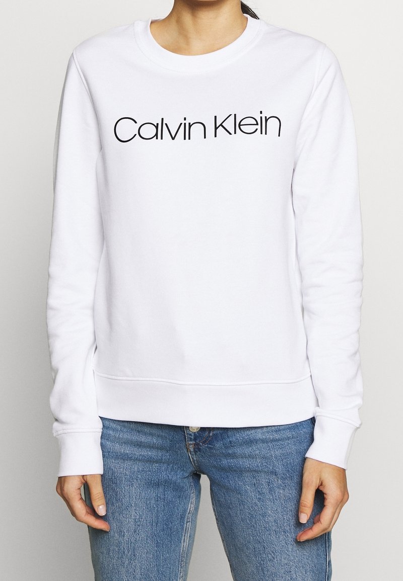 Kvinna klädd i vit långärmad sweatshirt från Calvin Klein och blåjeans, står med avslappnade händer vid sidorna mot en enfärgad bakgrund.