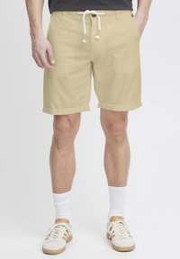Shorts beige con una texture liscia, orlo risvoltato, vita con cordoncino e un singolo bottone. Indossati con calzini bianchi e sneakers chiare.