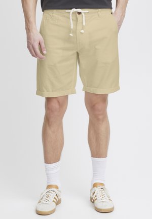 Shorts beige avec une texture lisse, ourlet retroussé, taille avec cordon de serrage et un seul bouton. Portés avec des chaussettes blanches et des baskets claires.