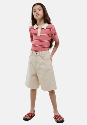 Fille debout, les mains dans le dos, portant un polo rose à rayures rouges, un short beige au genou et des sandales rouges et noires sur fond blanc.