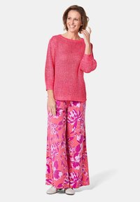Rosa gestrickter Pullover mit Dreiviertelärmeln, kombiniert mit weit geschnittenen Blumenhosen in Rosa- und Weißtönen. Getragen mit hellen Schuhen.