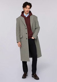 Světle šedý vlněný kabát s klopami s výřezem, zapínáním na jeden řádek knoflíků, postranní kapsy, vrstvený přes vínovou mikinu s kapucí a černé kalhoty.