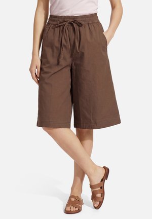 Vrouw draagt losse bruine knielange shorts met koord in de taille en bruine open teen platte sandalen, staat met één been gekruist.