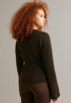 Jeune personne aux cheveux roux bouclés et volumineux portant un pull sombre en matière douce et un pantalon marron ajusté, debout de profil sur un fond neutre.