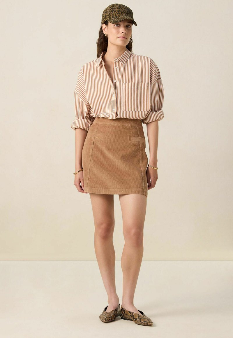 Brown corduroy mini skirt with front pockets, paired with a beige and white striped button-up shirt and a leopard print cap.