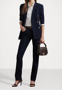 Kvinna med navyblå kavaj med guldknappar, vit skjorta, mörka jeans, brun bälte, högklackade sandaler och håller en liten brun handväska.