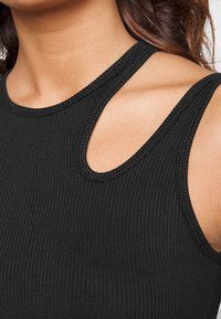 Närbild av en person som bär en svart ribbad ärmlös topp med en nyckelhålsklyvning nära halsringningen och axeln.