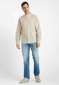 Man staat in een beige overhemd met mandarin kraag, lichtblauwe spijkerbroek en beige sneakers op een witte achtergrond.