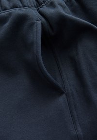 Tummansiniset jogger-housut, joissa on sileä pinta, joustava vyötärönauha ja sivutaskun avaus hienovaraisella ompelukoristeella.